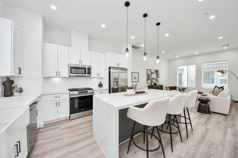 Bright, open-concept kitchen and living area with modern design. Features white cabinetry, stainless steel appliances, a large island with seating, and stylish pendant lighting. The space flows into a cozy living room with ample natural light.