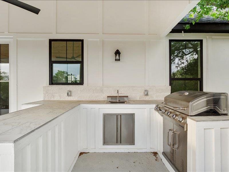 Outdoor kitchen featuring a built-in stainless steel grill, a sink with a faucet, and a refrigerator, all set within white cabinetry and light-colored countertops