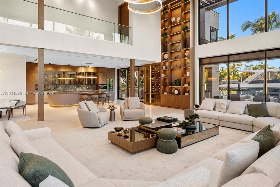 Wide-angle view of open-plan great room and chef's kitchen featuring curved fluted island with stone top, full-height illuminated walnut shelving, double-height glass walls with direct canal and yacht views, soaring ceilings, and light stone floors throughout.