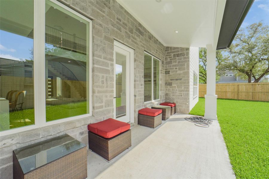 This photo features a spacious backyard patio with stone wall accents and large windows, offering a view of the lush green lawn. Comfortable seating with red cushions enhances the outdoor relaxation space.