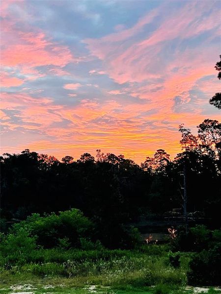 A view from the back of the home, the stunning sunrise sky with a palette of vibrant pinks and oranges above a tranquil, natural setting featuring lush trees and a glimpse of water, hinting at serene views from the property.