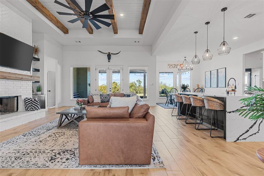 Living room featuring french doors, a brick fireplace, beamed ceiling, ceiling fan with notable chandelier, and light wood-type flooring