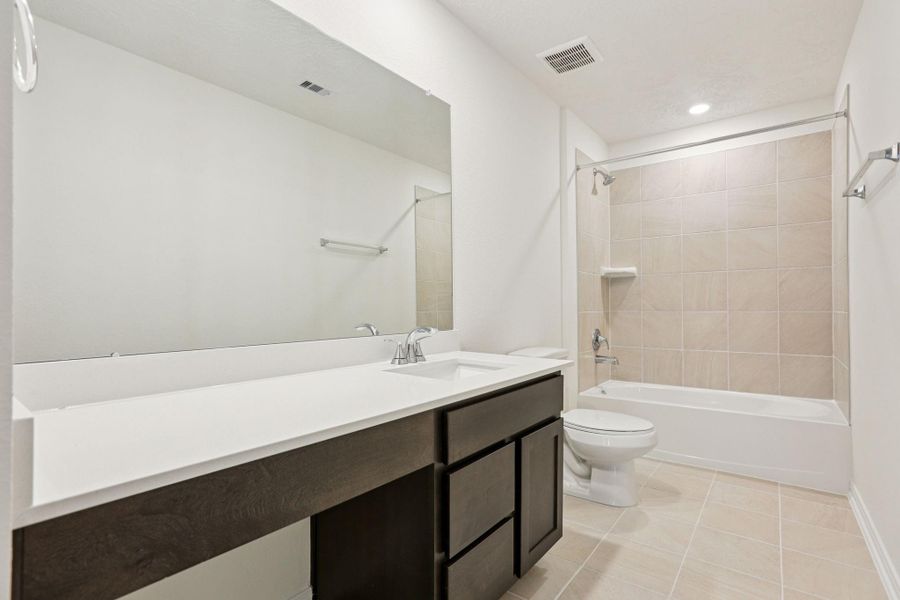 The secondary bathroom showcases tile flooring, a bath/shower combo with Daltile tile surround, a vanity with knee space and stunning light countertop, a mirror, sleek fixtures, and contemporary finishes throughout.