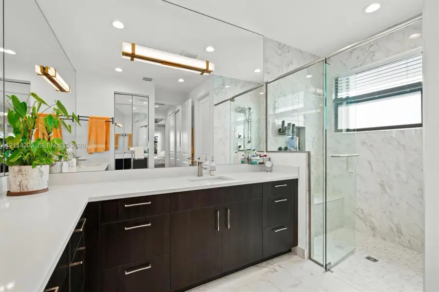 Bathroom featuring a dual-sink dark wood vanity with white countertops, a full-width mirror, recessed lighting, and a glass-enclosed shower with stone-tile surround