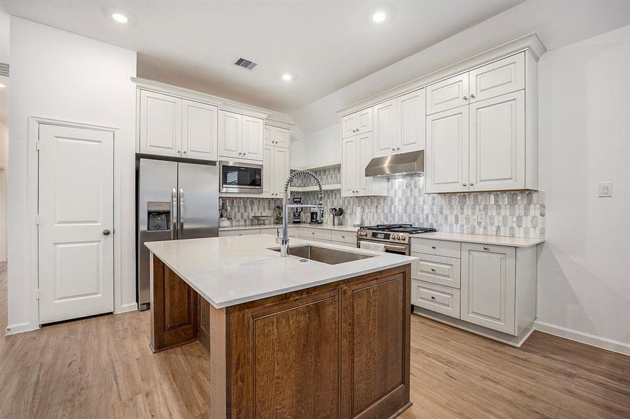 This modern kitchen features white cabinetry, stainless steel appliances, and a central island with a sleek countertop. The open layout flows into a dining area with ample natural light from a large window.