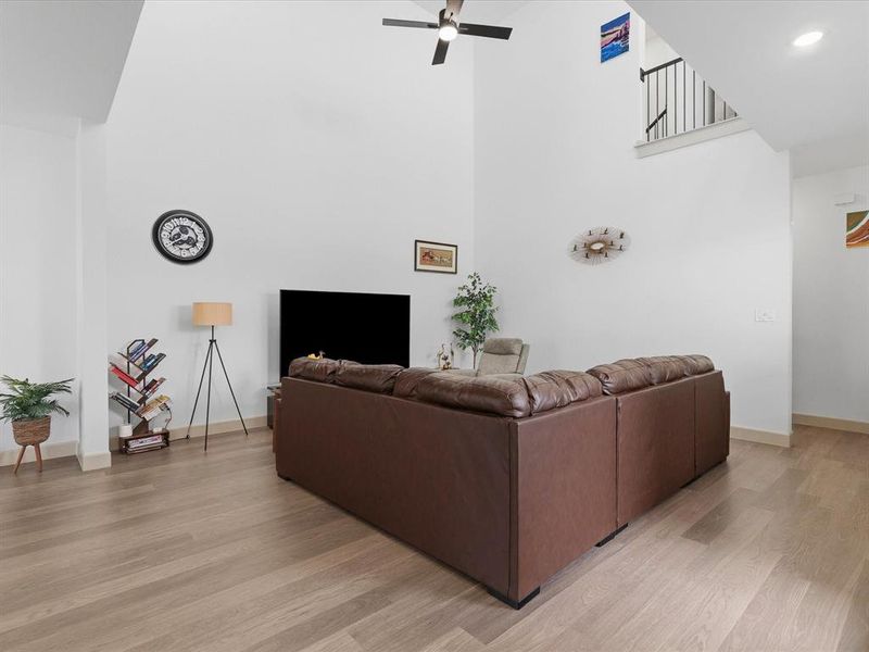 Spacious living area featuring a vaulted ceiling, ceiling fan, wood-finish flooring, white walls, and a staircase with black metal balusters