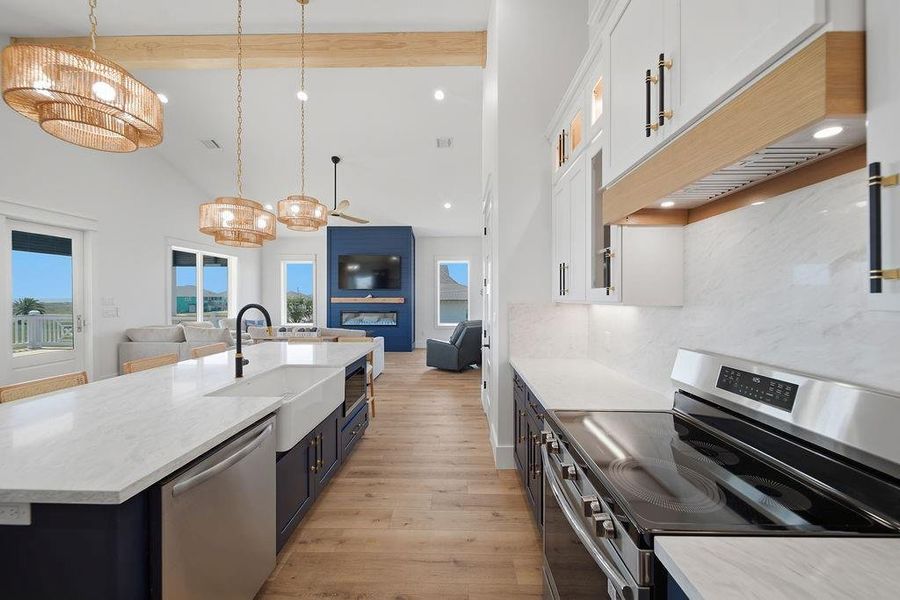 This bright, open-concept kitchen and living area features modern finishes, a large island with a farmhouse sink, sleek appliances, and stylish pendant lighting. The space flows into a cozy living room with a fireplace and large windows offering scenic views.
