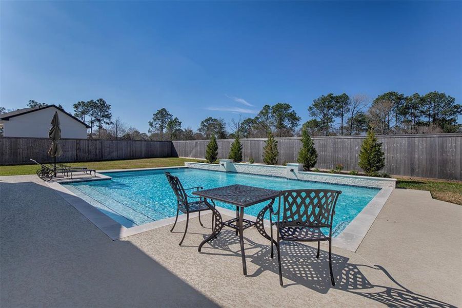 This backyard features a spacious in-ground pool with a surrounding patio, complete with outdoor furniture. It's enclosed by a wooden fence, providing privacy, and accented with a few small trees. Perfect for relaxation and entertaining.