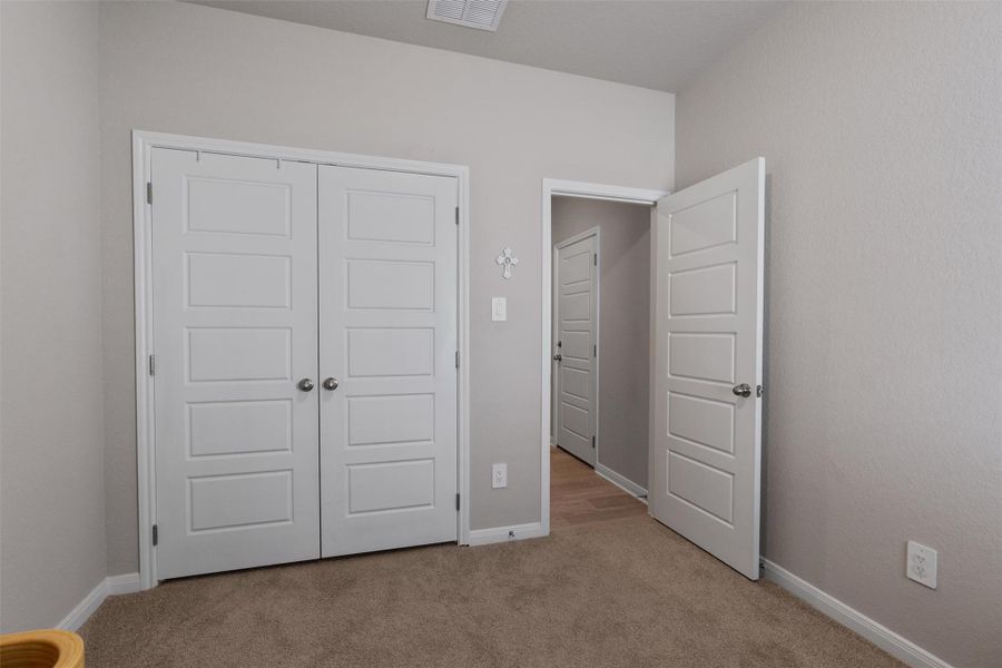 Neutral-toned room featuring a double-door closet with five-panel doors, light brown carpet, and an open doorway revealing a hallway with wood-finish flooring Neutral-toned room featuring a double-door closet with five-panel doors, light brown carpet, and an open doorway revealing a hallway with wood-finish flooring