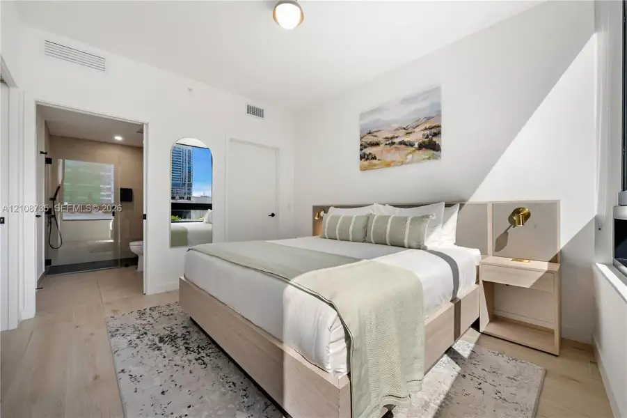 Bedroom featuring light wood-finish flooring, a built-in headboard with integrated lighting, and an arched wall mirror Bedroom featuring light wood-finish flooring, a built-in headboard with integrated lighting, and an arched wall mirror
