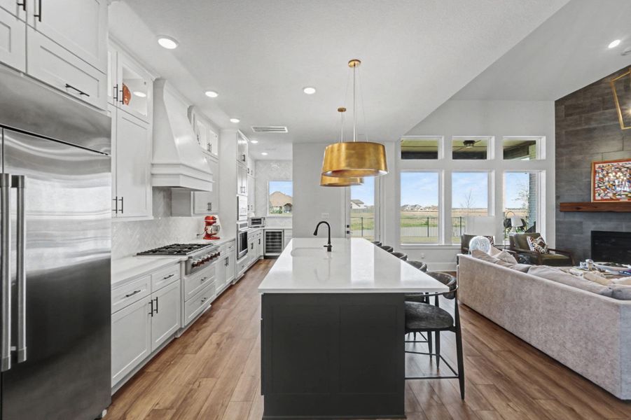 The heart of the kitchen features a professional-grade gas range and a substantial white range hood. Adjacent is a direct view into the functional butler’s pantry, equipped with a wine cooler and additional storage. Striking brass pendant lights hang over the oversized prep and dining island.