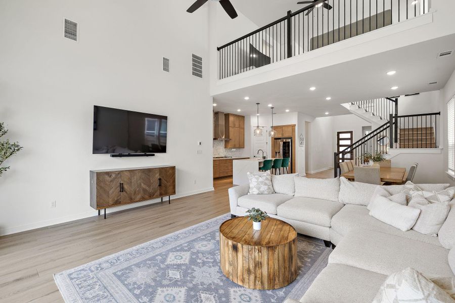 Open-concept living area featuring high ceilings, wood-finish flooring, a contemporary black metal staircase and railing, a kitchen visible with wooden cabinetry, and recessed lighting throughout