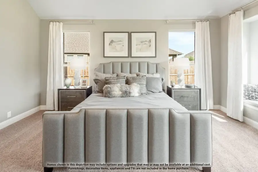 Bedroom with gray upholstered bed, carpet flooring, and two windows with white curtains