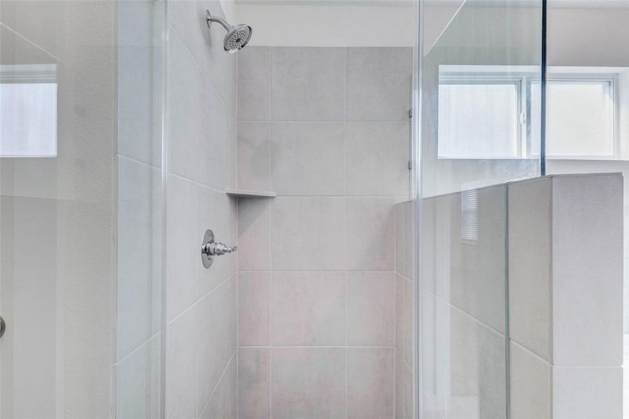 Close-up of the primary walk-in shower featuring neutral tile walls, glass enclosure, chrome fixtures, built-in corner shelf, and a frosted privacy window nearby. The shower’s clean design and bright finish create a fresh, low-maintenance bath feature with modern appeal.