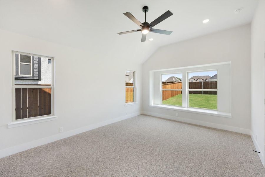 Empty bedroom with vaulted ceiling, ceiling fan, carpet flooring, and multiple windows overlooking backyard