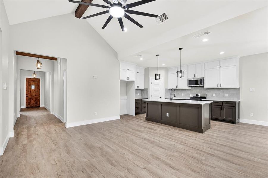 Spacious open-concept interior featuring light wood-finish flooring, vaulted ceiling with exposed beam and ceiling fan, and a modern kitchen with a large island, white shaker cabinetry, and stainless steel appliances