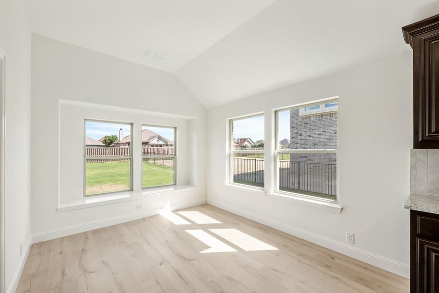 Empty room with vaulted ceiling, light wood floors, and large windows overlooking a backyard