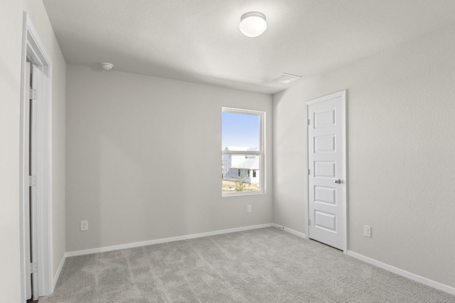 Image of a primary bedroom with tan carpeting, light grey walls, a white bathroom door and a window