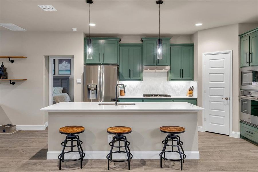 Kitchen with green cabinetry, light wood-style floors, stainless steel appliances, pendant lighting, and a kitchen island with sink