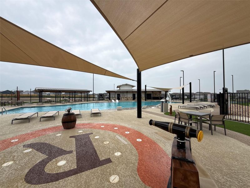 Expansive aquatic facility featuring a large swimming pool with integrated seating areas, multiple shade sails, and decorative ground surfacing