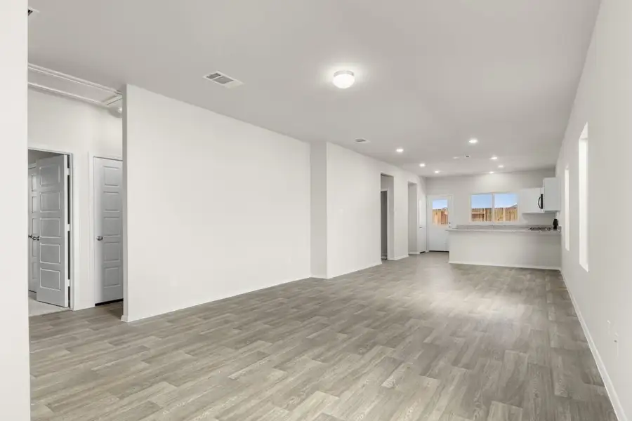 Image of home living spaces with wood-look flooring and light walls. Doorways and hallways in the left corner and kitchen in the back with multiple windows and door to the backyard Image of home living spaces with wood-look flooring and light walls. Doorways and hallways in the left corner and kitchen in the back with multiple windows and door to the backyard