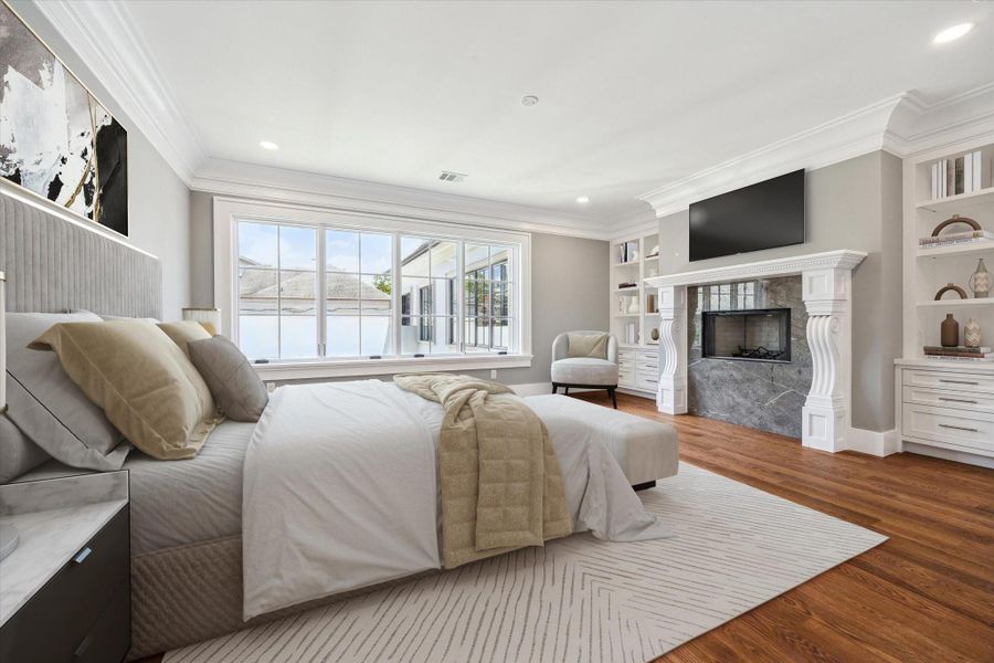 This primary bedroom features large windows for natural light, a cozy fireplace with black marble surround and custom mantle, built-in shelving, and elegant hardwood floors, beautiful crown molding and opens to the luxurious bath and closets.   This photo has been virtually staged.