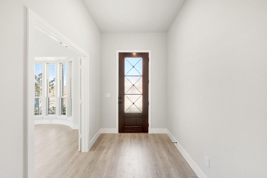 Entryway with dark wood glass-panel front door, light hardwood floors, and white walls leading to a bay window room. Entryway with dark wood glass-panel front door, light hardwood floors, and white walls leading to a bay window room.