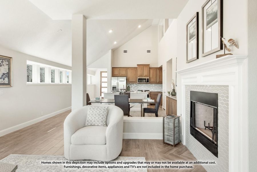 Open-concept living area with vaulted ceiling, white fireplace, and kitchen visible in background Open-concept living area with vaulted ceiling, white fireplace, and kitchen visible in background