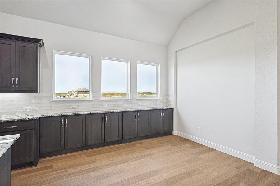Kitchen with tasteful backsplash, dark wood finish cabinets, light wood-style flooring, light stone countertops, and vaulted ceiling