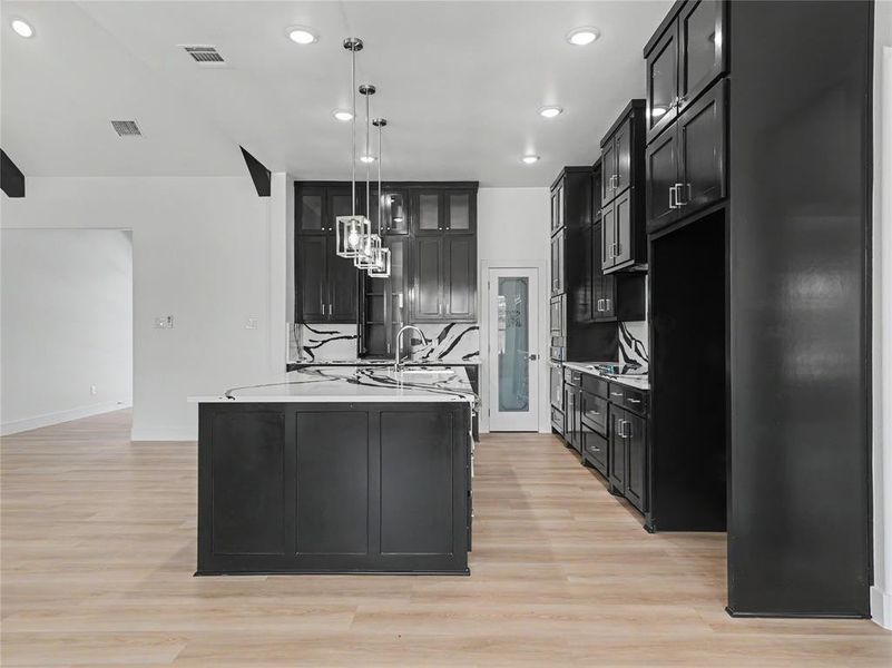 Kitchen featuring a central island with a sink, pendant lighting, dark cabinetry, light countertops, and wood-finish flooring Kitchen featuring a central island with a sink, pendant lighting, dark cabinetry, light countertops, and wood-finish flooring