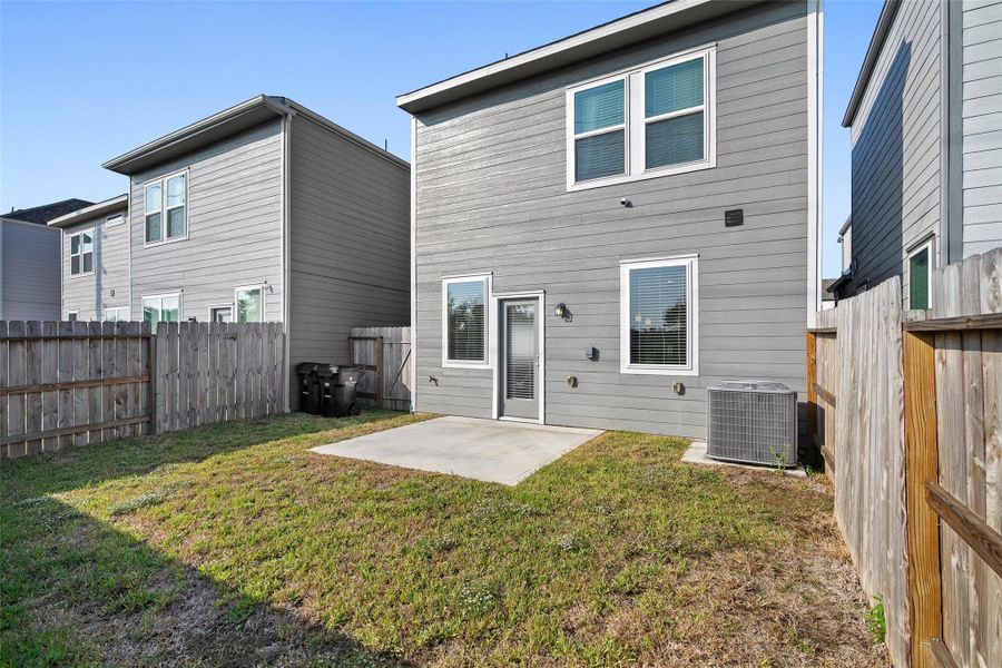 This photo shows the back of a modern two-story home with gray siding. It features a small fenced backyard with a concrete patio, ideal for outdoor activities or relaxation. The yard is bordered by wooden fences, providing privacy.