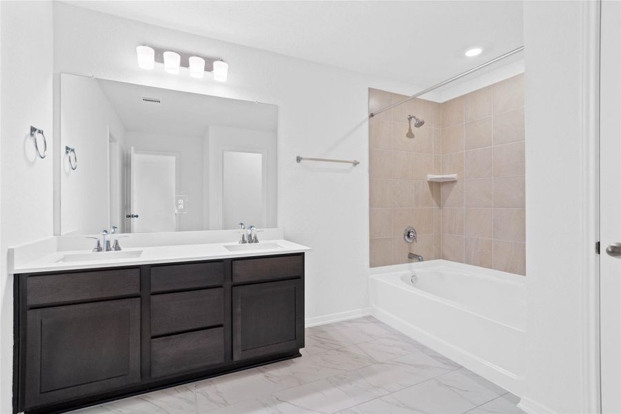 The primary bathroom offers a spacious tub/shower combination with a tiled surround, stained cabinetry topped with light countertops, dual sinks, high ceilings, neutral paint, a large mirror, and sleek, modern finishes for a polished look.