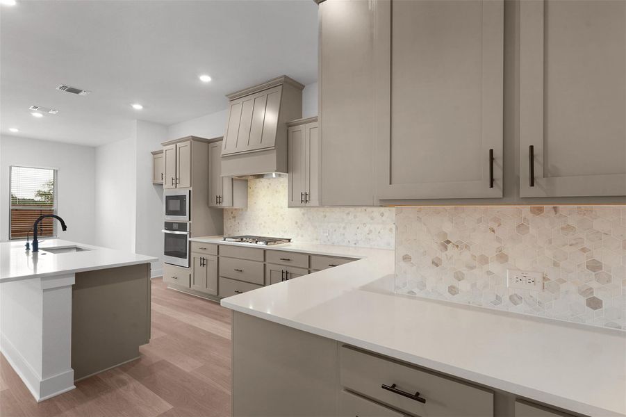 Kitchen featuring gray cabinets, decorative backsplash, light wood-style flooring, appliances with stainless steel finishes, and premium range hood