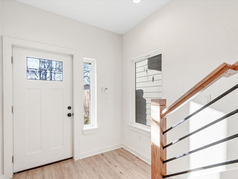 Bright entryway featuring a modern front door with frosted glass, window, and sleek wooden stairs with metal railing accents, all set against light walls and flooring.