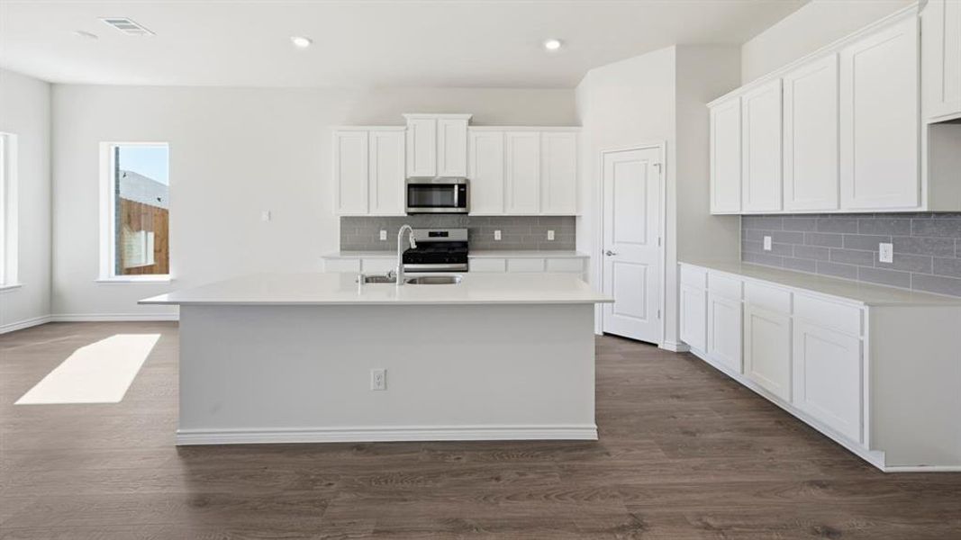 Open-concept kitchen featuring an island with an undermount sink, stainless steel appliances, white cabinetry, a subway tile backsplash, and wood-finish flooring