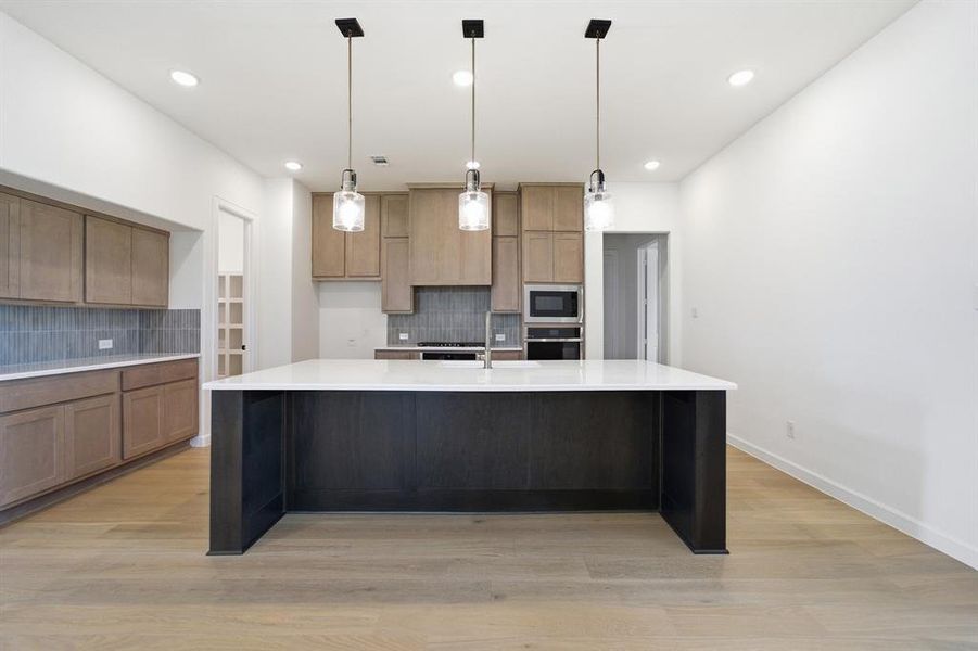 Spacious kitchen featuring a large central island with a white countertop and three pendant lights, light wood-finish cabinetry, built-in stainless steel appliances, and wood-finish flooring