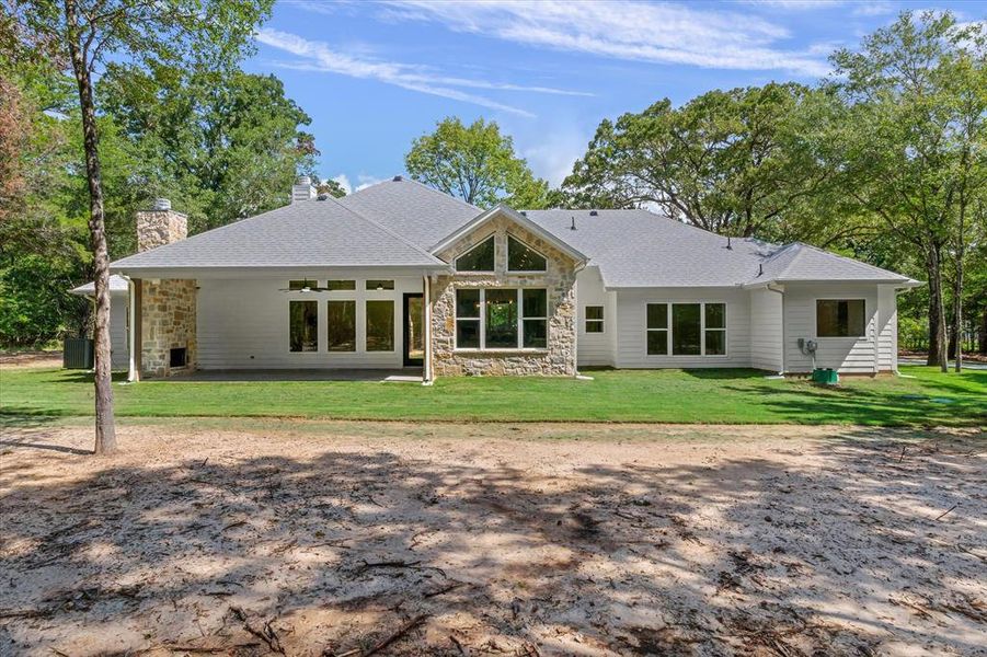 Back of house with stone siding, a patio area, a lawn, roof with shingles, and a chimney Back of house with stone siding, a patio area, a lawn, roof with shingles, and a chimney