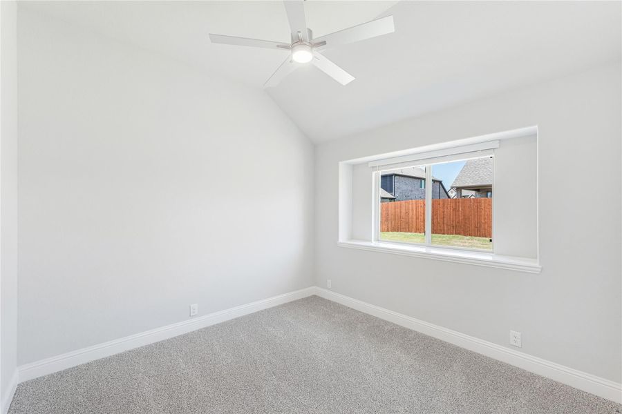 Empty bedroom with vaulted ceiling, ceiling fan, carpet flooring, and a window overlooking a backyard fence.