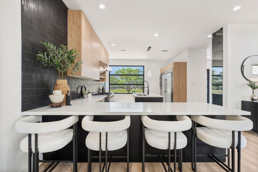 The kitchen island features a generous white countertops with seating along the breakfast bar side, framing a direct view of the kitchen's most striking design moment — warm natural wood upper cabinetry set against a vertical black tile backsplash. The contrast is intentional and it lands.
