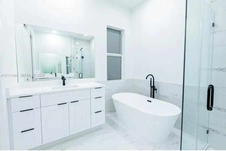 Freestanding soaking tub with matte black floor-mounted faucet, white cabinetry with quartz countertop, integrated rectangular sink, large wall-mounted mirror, and glass-enclosed shower with tiled surround Freestanding soaking tub with matte black floor-mounted faucet, white cabinetry with quartz countertop, integrated rectangular sink, large wall-mounted mirror, and glass-enclosed shower with tiled surround