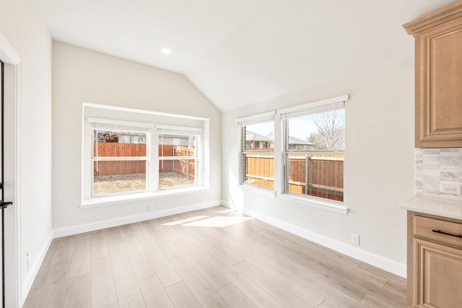 Bright empty dining area with vaulted ceiling, light wood floors, and large windows overlooking backyard fence Bright empty dining area with vaulted ceiling, light wood floors, and large windows overlooking backyard fence