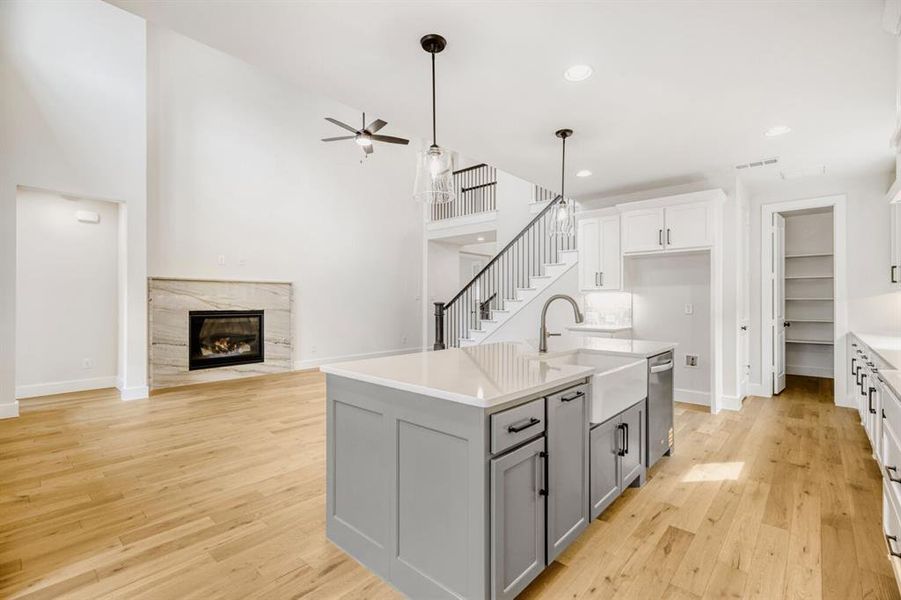 Open-concept kitchen and living area featuring a central island with a farmhouse sink, light wood-finish flooring, a contemporary fireplace with a stone surround, and a staircase with dark railings