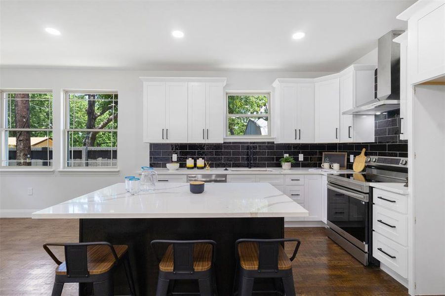 Kitchen with stainless steel range with electric stovetop, wall chimney range hood, a breakfast bar, backsplash, and recessed lighting Kitchen with stainless steel range with electric stovetop, wall chimney range hood, a breakfast bar, backsplash, and recessed lighting