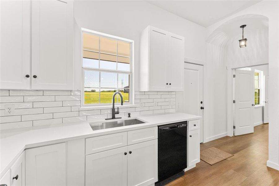 Kitchen featuring white shaker-style cabinetry, white subway tile backsplash, stainless steel double-basin sink with a matte black gooseneck faucet, white solid surface countertops, and wood-finish flooring
