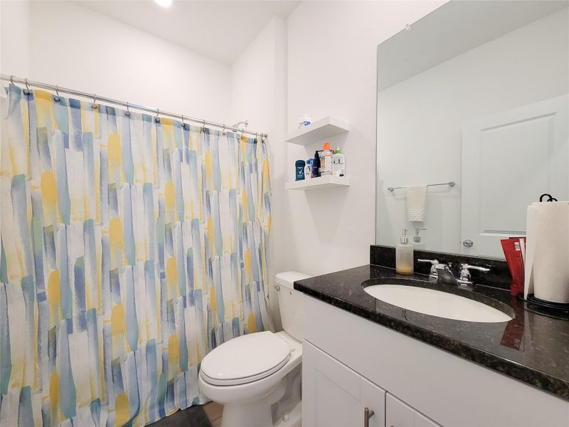This secondary bathroom features a modern design with a sleek black countertop, white cabinetry, and a large mirror. It includes a shower and shelving for storage, providing both functionality and style.