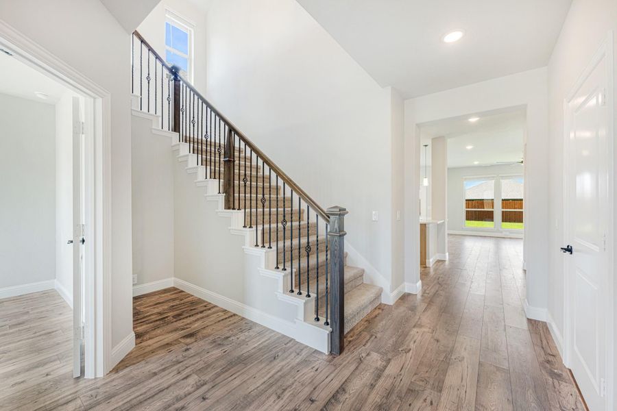 Bright entryway with carpeted staircase, iron balusters, wood handrail, and hardwood floors leading to open living area