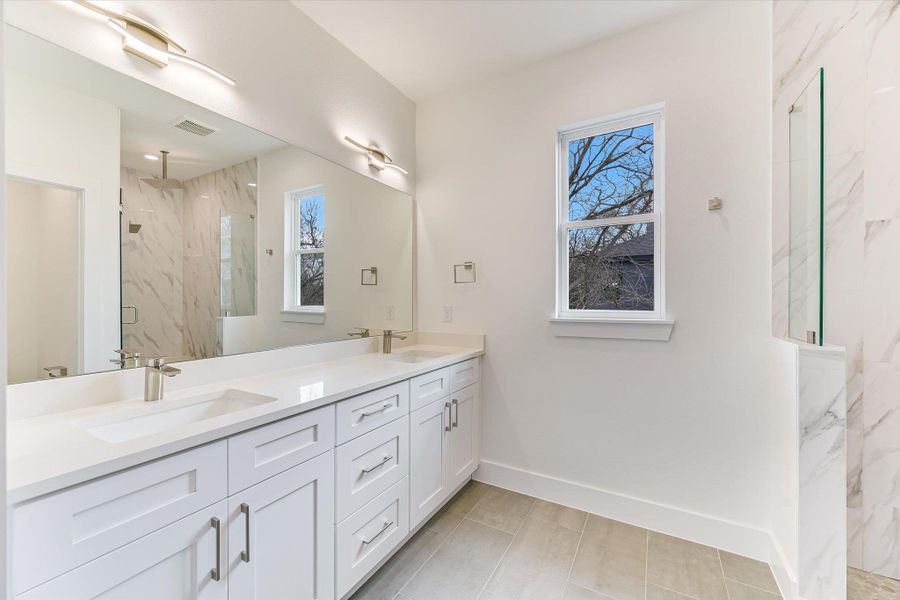 Step into a spa-like retreat featuring a sleek double vanity with crisp white shaker cabinetry and modern brushed nickel hardware. The expansive mirror and designer lighting create a bright, airy atmosphere, while the walk-in shower showcases elegant marble-patterned tiling.