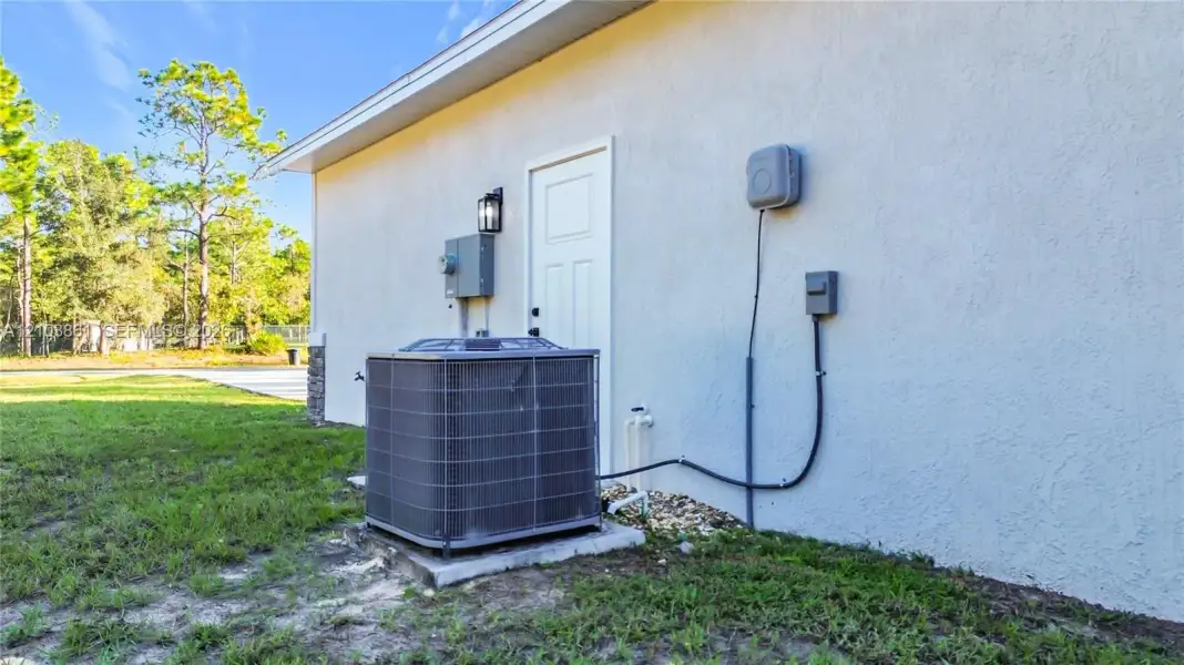 Exterior stucco wall featuring a single-entry door, external wall-mounted lighting, and a central heating and cooling unit on a concrete pad
