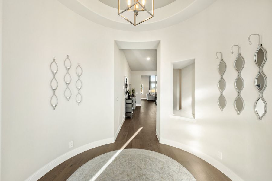 Round foyer with tray ceiling, geometric chandelier, hardwood floors, and decorative wall mirrors leading to hallway Round foyer with tray ceiling, geometric chandelier, hardwood floors, and decorative wall mirrors leading to hallway