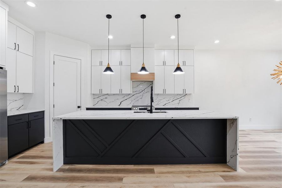 Spacious kitchen featuring a waterfall island with decorative paneling, white shaker cabinetry, black base cabinets, a built-in sink with a black faucet, and light wood-finish flooring
